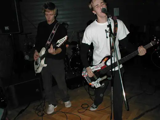 The punk band on stage at secondary school in 2003: a guitarist, a singer on bass, and a drummer behind them.