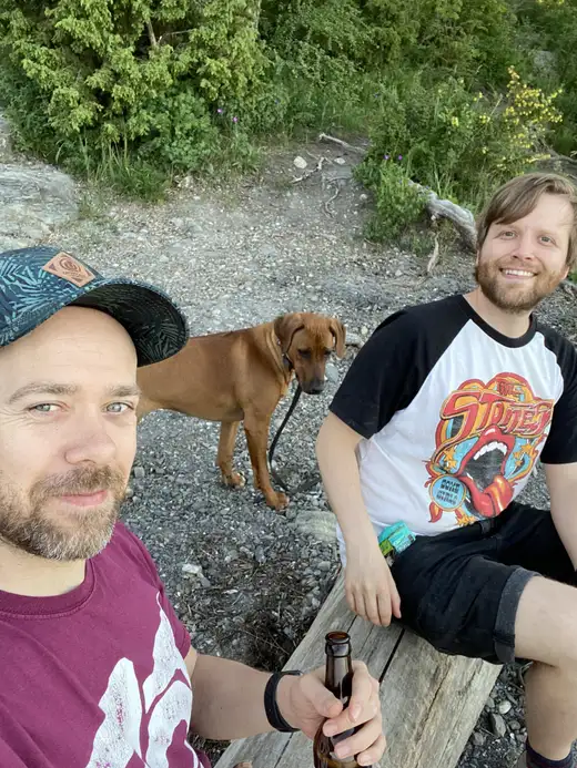 Kristoffer in a cap and Finn on a bench by the shore, a dog standing behind them.