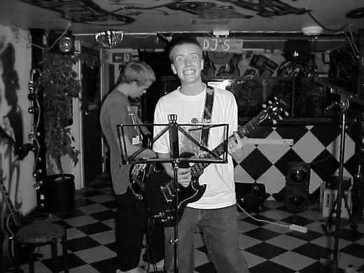 A rehearsal in 2001 at a club with a chequered floor: a guitarist grinning at the camera, another guitarist behind.